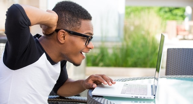 Smiling preppy guy in glasses leans over laptop, holding a hand to the back of his head