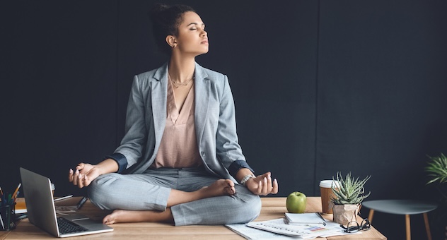 Person in business suit and blouse sits in meditation in lotus pose on top of desk alongside laptop, cactus pot plant, takeaway cup and documents