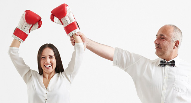 A woman stands to the left of frame with boxing gloves on her hands. She is raising her gloved hands over her head in triumph. A man in a white shirt and bowtie stands next to her.