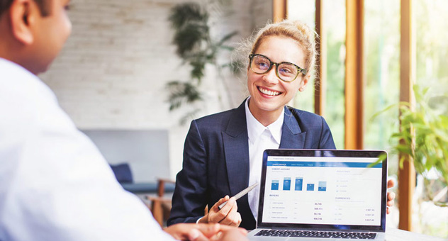 A business woman sits at a desk, presenting a proposal (on a laptop screen) to the man sitting across from her.