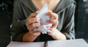 A woman sitting at a desk crumpling white paper between her hands.