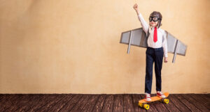 A little boy wearing cardboard wings. He is standing on a skateboard with his hand in the air looking triumphant.