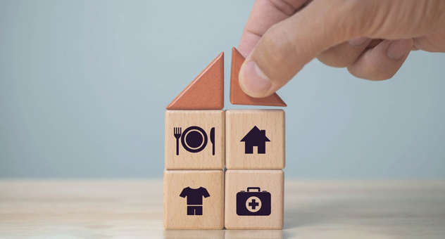 Hand stacks wooden blocks with icons of house, plate and cutlery, clothes and first aid kit