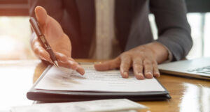 Close-up of a man sitting at a desk. The desk has papers on it and the man is reaching out a hand in an inviting way - as if offering up information.