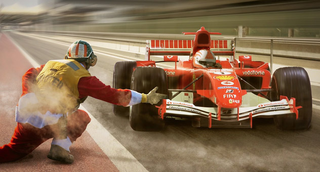 A red race car stopped briefly on a racetrack. A pit crew man in a red boilersuit with a yellow vest flags the car down.