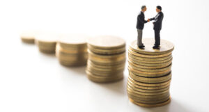 Two miniature, plastic toy business men shaking hands on top of a stack of coins.