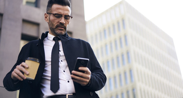 A man in business attire with a smartphone in one hand and a coffee in the other.