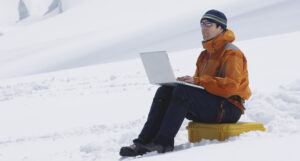 A man in winter gear sitting on a snowy slope with a laptop on his knees.