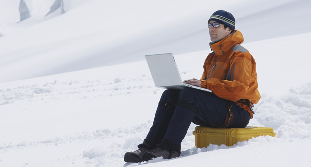 A man in winter gear sitting on a snowy slope with a laptop on his knees.