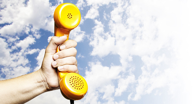 A hand holding a yellow phone receiver / handset against a blue sky.