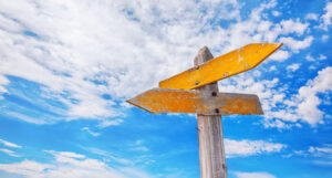 Two yellow arrow-shaped signs on a post against a blue sky.