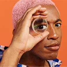 A man with pink hair looks at the camera with a lens held to his eye.